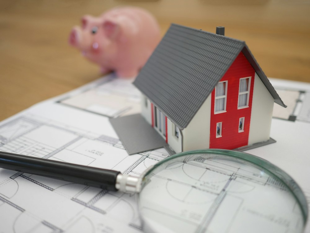 Miniature house on architectural blueprints with a piggy bank in the background, illustrating financial planning for real estate investment.
