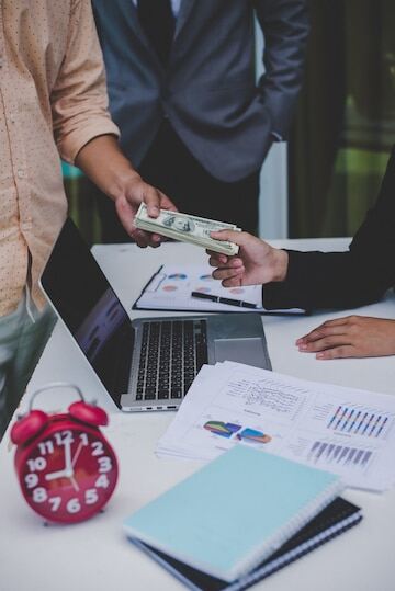 An image of a person giving loan to another with a laptop, alarm clock, and papers on the desk