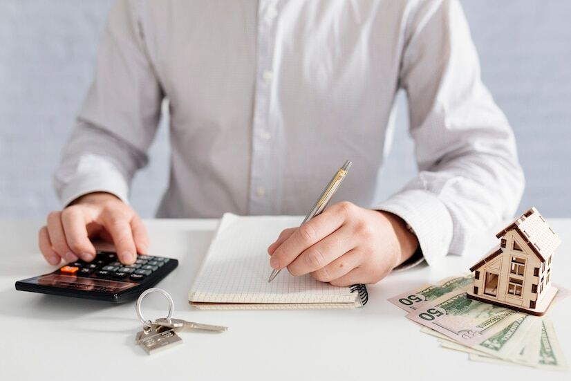 A person writing on a notepad while using a calculator with a house key, home model, and dollar notes on the table