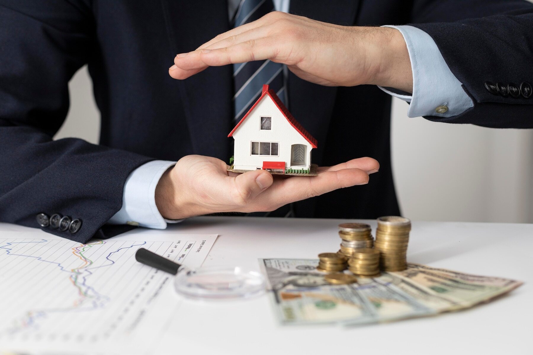 A hard money lender holding a miniature house with cash and coins on the table