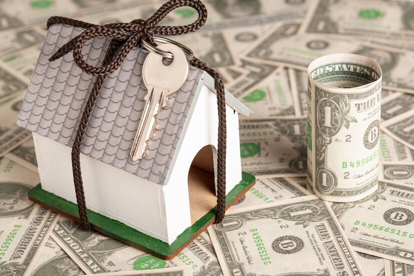 An image of a miniature house tied with a key and rope surrounded by US dollar banknotes